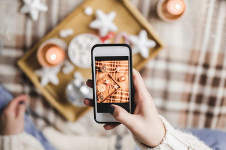 Female hand with smartphone takes pictures of New Years or Christmas flatlay for social networks. Preparing for holidays, creating content. Wooden tray with cocoa mug marshmallows, candle, stars.の写真素材