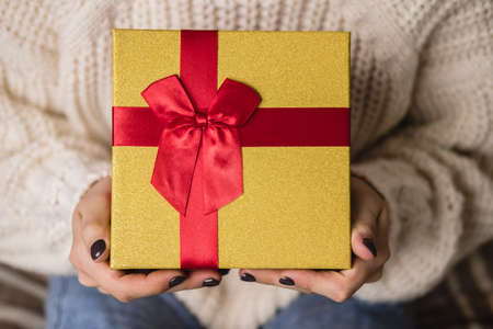 Young woman sits on bed in cozy knitted wool white sweater and holding gift-box with red gold....