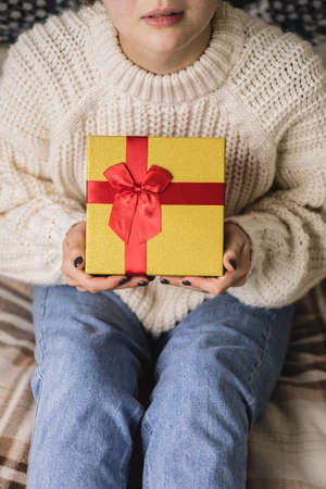 Young woman sits on bed in cozy knitted wool white sweater and holding gift-box with red gold. Hygge, New Years, Christmas, holiday preparations. Candles, Christmas balls, gifts, cocoa marshmallows.の写真素材