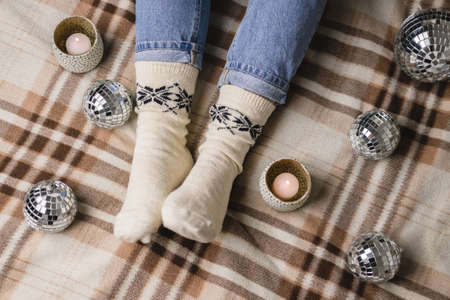 Young woman is sitting on the bed in cozy knitted woolen white sweater, socks and holding toy disco ball in her hands. Hygge, candles, New Years, Christmas, holiday preparations.の写真素材