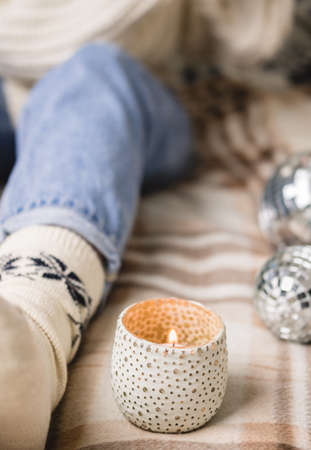 Young woman is sitting on the bed in cozy knitted woolen white sweater, socks and holding toy disco ball in her hands. Hygge, candles, New Years, Christmas, holiday preparations.の写真素材