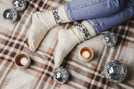 Young woman is sitting on the bed in cozy knitted woolen white sweater, socks and holding toy disco ball in her hands. Hygge, candles, New Years, Christmas, holiday preparations.の写真素材