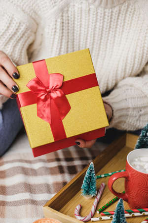 Young woman sits on bed in cozy knitted wool white sweater and holding gift-box with red gold. Hygge, New Years, Christmas. Wooden tray with a mug of cocoa with marshmallows, toy tree, candle, stars.の写真素材