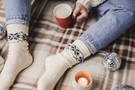 Young woman sits on plaid in cozy knitted woolen white sweater and socks holds cup of cocoa in her hands. Hygge New Year, cozy Christmas, preparation for holidays. Candles, Christmas balls.の写真素材