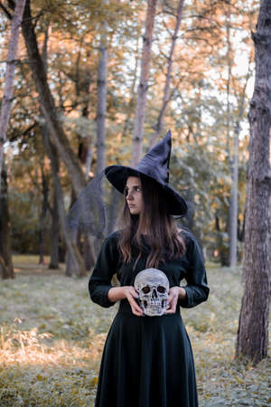 Young beautiful cute woman in dark dress and witchs hat holds a skull in her hands. Halloween party costume. Forest, park with autumn trees.の写真素材