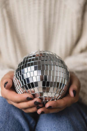 Young woman is sitting on the bed in cozy knitted woolen white sweater, socks and holding toy disco ball in her hands. Hygge, New Years, Christmas, holiday preparations.の写真素材