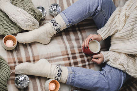 Young woman sits on plaid in cozy knitted woolen white sweater and socks holds cup of cocoa in her hands. Hygge New Year, cozy Christmas, preparation for holidays. Candles, Christmas balls.の写真素材