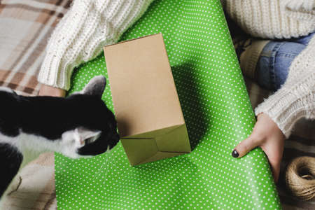 Young woman sits on plaid in cozy knitted woolen white sweater, socks and wraps Christmas gift in...