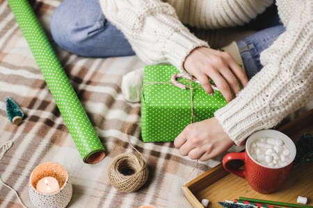 Young woman sits on plaid in cozy knitted woolen white sweater, socks and wraps Christmas gift in polka dot wrapping paper. Wooden tray with mug of cocoa with marshmallows, toy tree, candle, straws.の写真素材