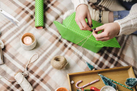 Young woman sits on plaid in cozy knitted woolen white sweater, socks and wraps Christmas gift in polka dot wrapping paper.の写真素材
