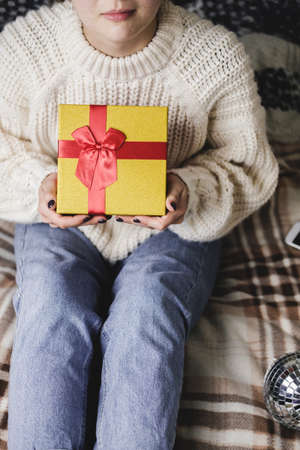 Young woman sits on bed in cozy knitted wool white sweater and holding gift-box with red gold. Hygge, New Years, Christmas, holiday preparations. Candles, Christmas balls, gifts, cocoa marshmallows.の写真素材