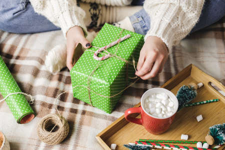 Young woman sits on plaid in cozy knitted woolen white sweater, socks and wraps Christmas gift in polka dot wrapping paper. Wooden tray with mug of cocoa with marshmallows, toy tree, candle, straws.の写真素材