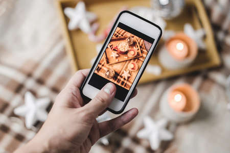 Female hand with smartphone takes pictures of New Year's or Christmas flatlay for social networks. Preparing for holidays, creating content. Wooden tray with cocoa mug marshmallows, candle, starsの写真素材