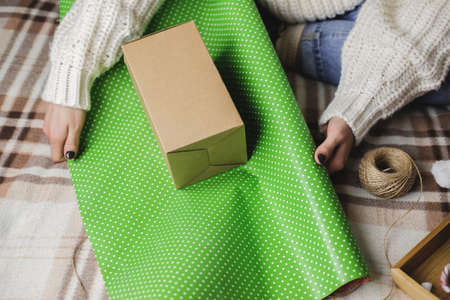 Young woman sits on plaid in cozy knitted woolen white sweater, socks and wraps Christmas gift in...
