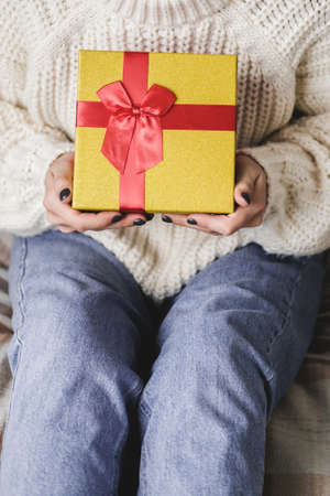 Young woman sits on bed in cozy knitted wool white sweater and holding gift-box with red gold. Hygge, New Years, Christmas, holiday preparations. Candles, Christmas balls, gifts, cocoa marshmallowsの写真素材