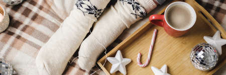 Young woman sits on plaid in cozy knitted woolen white sweater and holds cup of cocoa with marshmallows. Hygge banner New Year, cozy preparation for holidays. Candles, Christmas balls in wooden trayの写真素材