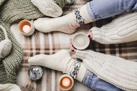 Young woman sits on plaid in cozy knitted woolen white sweater and socks holds cup of cocoa in her hands. Hygge New Year, cozy Christmas, preparation for holidays. Candles, Christmas ballsの写真素材