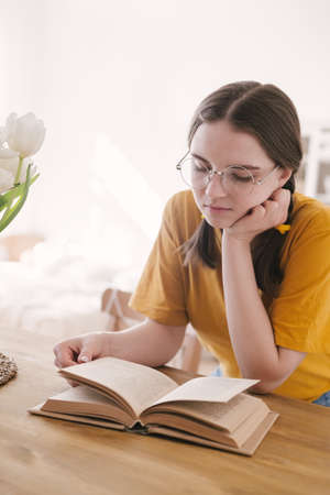 Young beautiful woman student in orange T-shirt and glasses reads book sitting at the table....