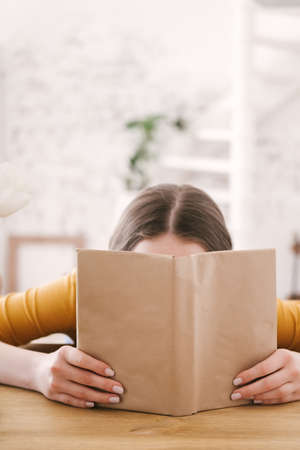 Young beautiful woman student in orange T-shirt and glasses reads book sitting at the table. Preparing for exams, studying at home on self-isolation.の写真素材