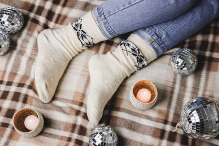 Young woman is sitting on the bed in cozy knitted woolen white sweater, socks and holding toy disco ball in her hands. Hygge, candles, New Years, Christmas, holiday preparations.の写真素材
