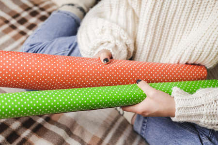 Young woman sits on plaid in cozy knitted woolen white sweater, socks and wraps Christmas gift in polka dot wrapping paper. Wooden tray with mug of cocoa with marshmallows, toy tree, candle, straws.の写真素材