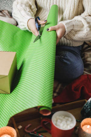 Young woman sits on plaid in cozy knitted woolen white sweater, socks and wraps Christmas gift in polka dot wrapping paper. Wooden tray with mug of cocoa with marshmallows, toy tree, candle, straws.の写真素材