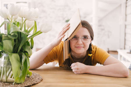 Young beautiful woman student in orange T-shirt and glasses reads book sitting at the table. Preparing for exams, studying at home on self-isolation.の写真素材