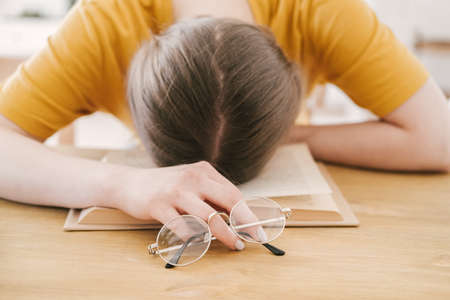 Young beautiful woman student in orange T-shirt and glasses reads book sitting at the table. Preparing for exams, studying at home on self-isolation.の写真素材