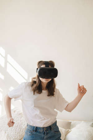 Young woman in jeans and white T-shirt with virtual reality glasses on her head moves her arms, dances, gets scared. The concept of modern technology, VR, virtual games, entertainment.の写真素材