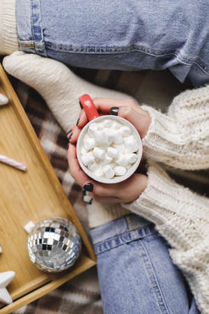 Young woman sits on plaid in cozy knitted woolen white sweater and holds cup of cocoa with marshmallows.の写真素材