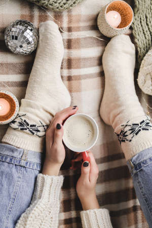 Young woman sits on plaid in cozy knitted woolen white sweater and socks holds cup of cocoa in her hands.の写真素材