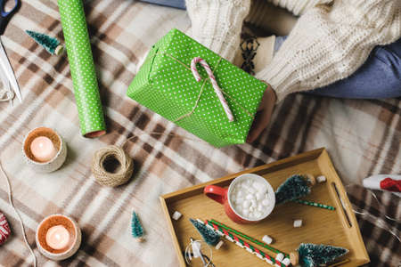 Young woman sits on plaid in cozy knitted woolen white sweater, socks and wraps Christmas gift in polka dot wrapping paper.の写真素材