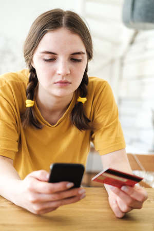 Cute young woman in bright yellow T-shirt sits at table and makes purchases online using smartphone and credit card. Shopping online during the coronavirus pandemic.の写真素材