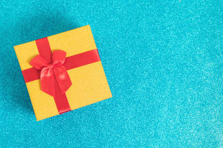 Golden glittering gift box withred bow on sparkling blue glitter background. Christmas and New Year background for congratulations on the coming holidays. Gift exchange. Flat lay. Top view. Copyspace.の写真素材