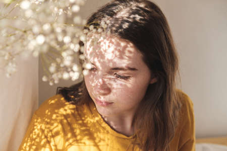 Portrait of young beautiful woman by the window with shadow from flowers on her face. Morning spring...