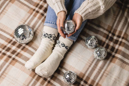 Young woman is sitting on the bed in cozy knitted woolen white sweater, socks and holding toy disco ball in her hands. Hygge, New Years, Christmas, holiday preparations.の写真素材