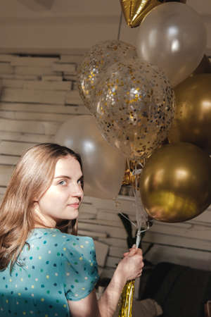 Young beautiful girl in blue dress with white polka dots celebrates her birthday and enjoys the golden balloons. Birthday alone at home during self-isolation.の写真素材