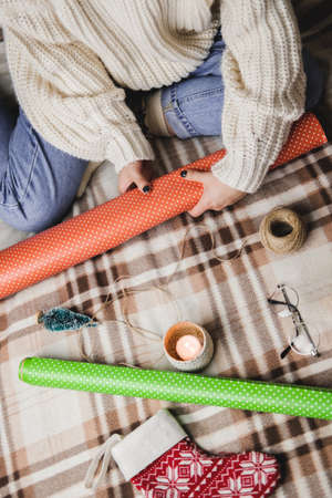 Young woman sits on plaid in cozy knitted woolen white sweater, socks and wraps Christmas gift in polka dot wrapping paper. Wooden tray with mug of cocoa with marshmallows, toy tree, candle, straws.の写真素材