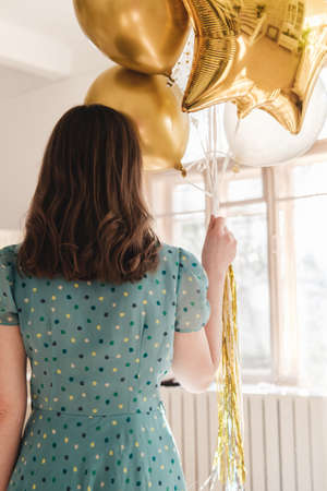 Young beautiful girl in blue dress with white polka dots celebrates her birthday and enjoys the golden balloons. Birthday alone at home during self-isolation.の写真素材