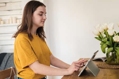 Young beautiful woman in yellow t-shirt usimg tablet for work, online business, studying sitting at home. Self-isolation, study, work during quarantine, pandemic. Remote job, education, student.の写真素材