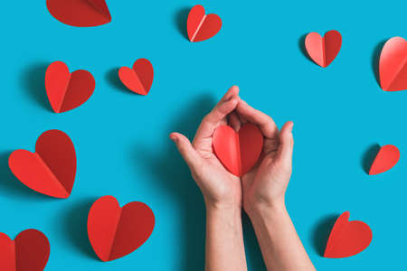 Festive background with red paper hearts for celebration of Valentines Day. Womens hands hold the heart. Flatlay by 14 February on bright blue background. Top view.の写真素材