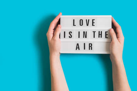 Festive background with woman hands holding glowing lightbox with text Love is in the Air on bright blue background for Valentines Day. Flatlay for 14th February. Top view. Copyspace.の写真素材