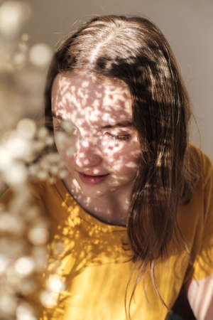 Portrait of young beautiful woman by the window with shadow from flowers on her face. Morning spring aesthetics.の写真素材