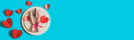 Banner with served table with plate and cutlery forcelebration of Valentines Day. On plate is napkin with paper heart. Flatlay website banner on bright blue background with red hearts. Top view.の写真素材