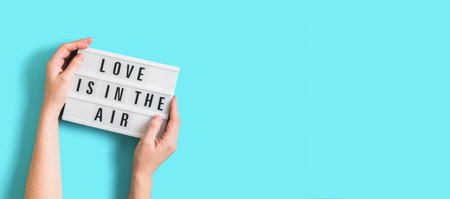 Festive banner with woman hands holding glowing lightbox with text Love is in the Air on bright blue background for Valentines Day. Flatlay banner for 14th February. Top view. Copyspace.の写真素材