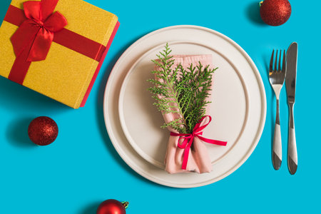 Served table with plate and cutlery for celebration of Christmas and New Year. On plate is napkin with a Christmas tree branch, red balls. Flatlay on blue background with balls, gift box.の写真素材