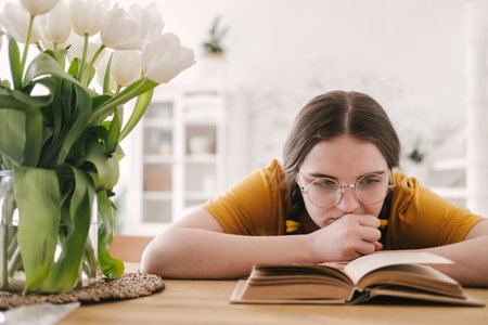 Young beautiful woman student in orange T-shirt and glasses reads book sitting at the table. Preparing for exams, studying at home on self-isolation.の写真素材