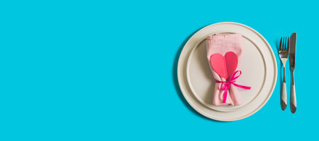 Banner with served table with plate and cutlery for the celebration of Valentines Day. On the plate is napkin with paper heart. Flatlay website banner on bright blue background. Top view.の写真素材
