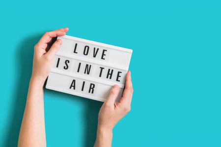 Festive background with woman hands holding glowing lightbox with text Love is in the Air on bright...