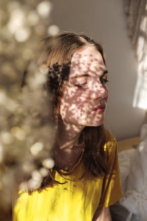 Portrait of young beautiful woman in yellow illuminating t-shirt by the window with grey shadow from flowers on her face. Morning spring aesthetics.の写真素材